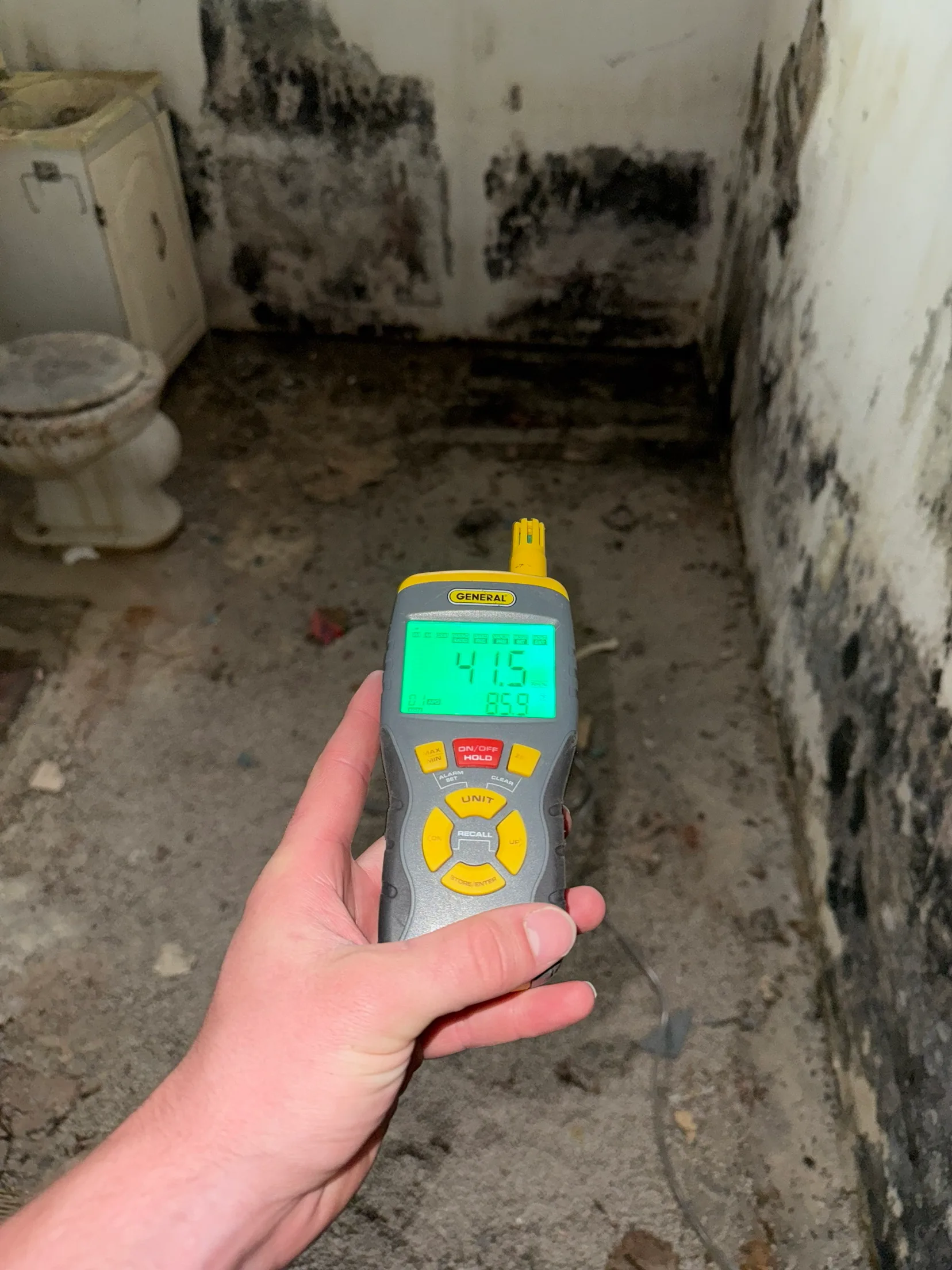 A thermo-hygrometer reading 41.5 percent relative humidity at 85.9 degrees, held up in a bathroom where black mold covers the lower walls and the floor has been stripped back to the slab.